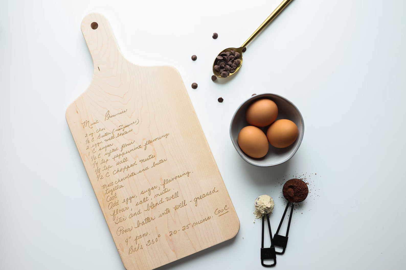 Wooden cutting board with handwritten recipe, bowl of eggs, and kitchen utensils on a white background